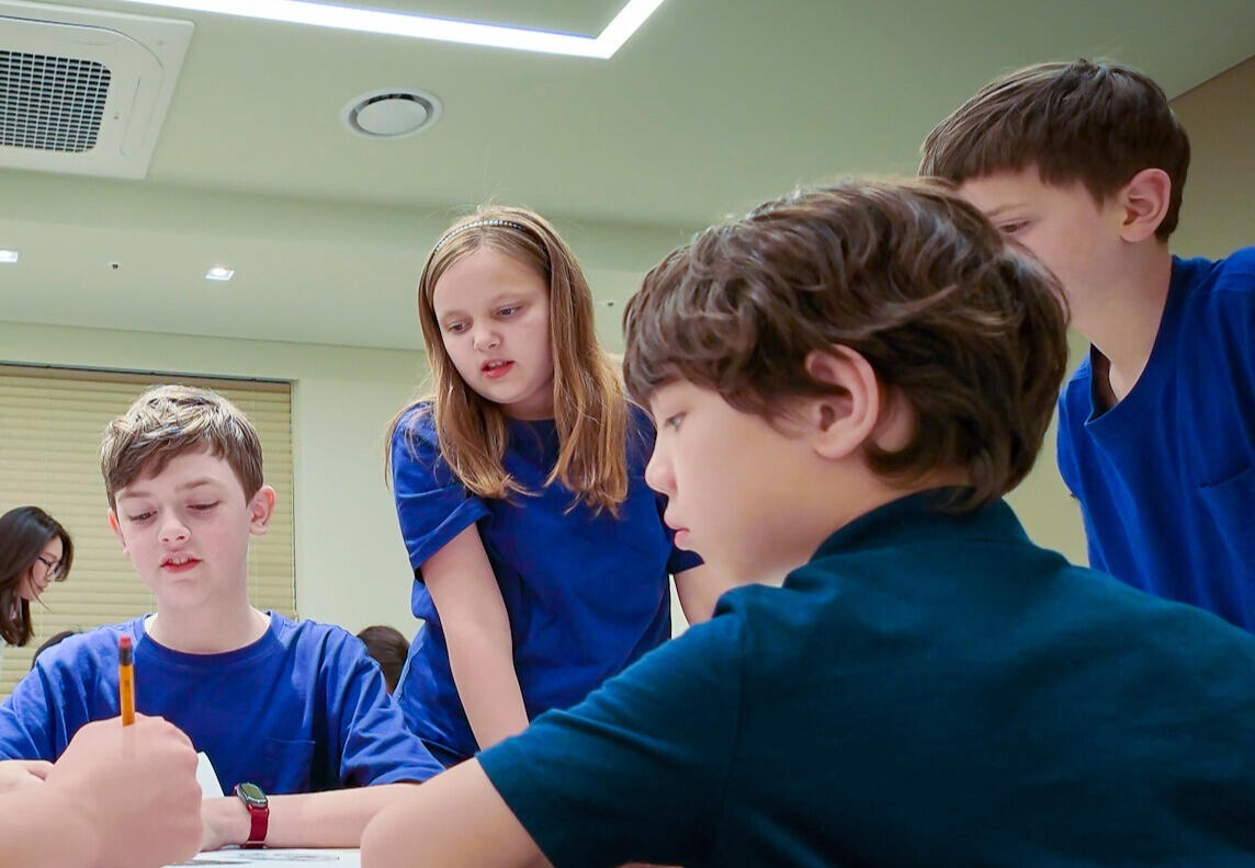A group of expats wearing aprons and gloves participate in an outdoor Korean kimchi-making event, handling napa cabbage and red pepper paste on a large preparation table.